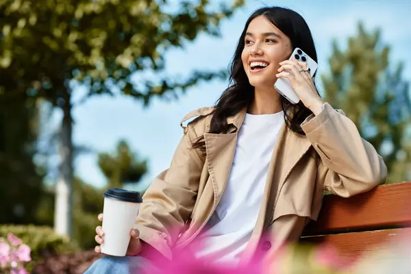 In the park, a young woman dressed warmly chats happily on her phone while enjoying coffee. — Stock Photo