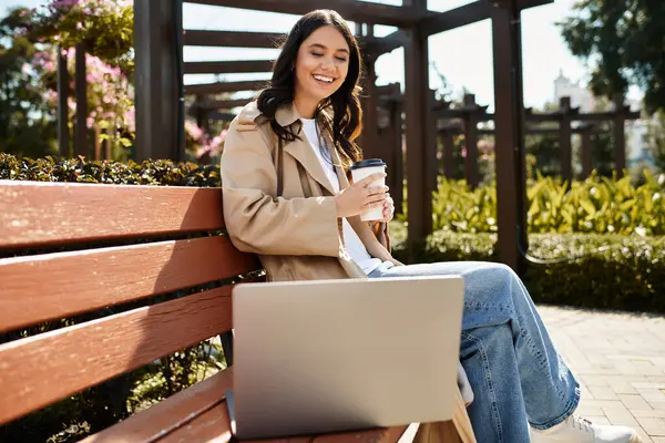 A young woman sits on a bench in the park, relaxing with coffee and focused on her laptop. — Stock Photo