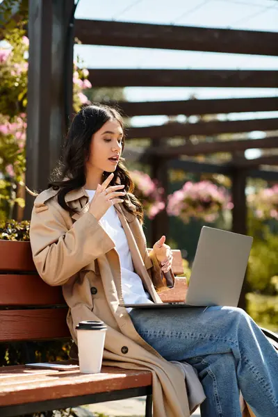 A young woman in warm attire sips coffee while using her laptop in a peaceful park. — Stock Photo