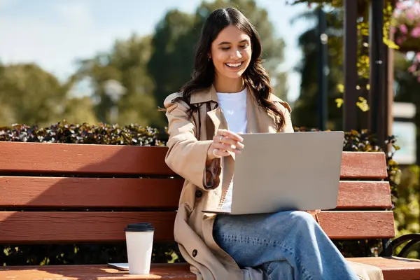 Vestida con un atuendo cálido, una mujer sonríe mientras trabaja en su computadora portátil en un parque de otoño. - foto de stock