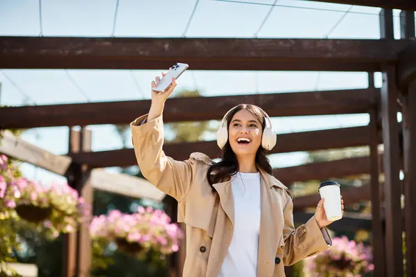 Una donna allegra indossa accoglienti danze autunnali nel parco, tenendo il telefono e il caffè. — Foto stock