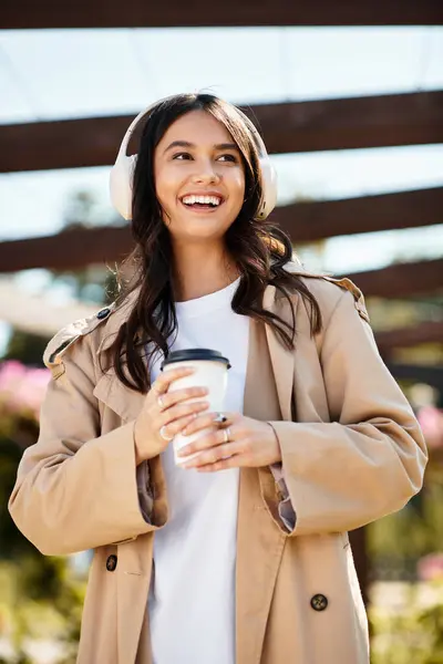 The cheerful woman in warm attire strolls through a vibrant park, sipping her drink and smiling. — Stock Photo