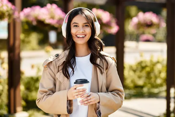 Una mujer alegre en traje de otoño acogedor sonríe mientras sostiene una taza de café al aire libre. — Stock Photo