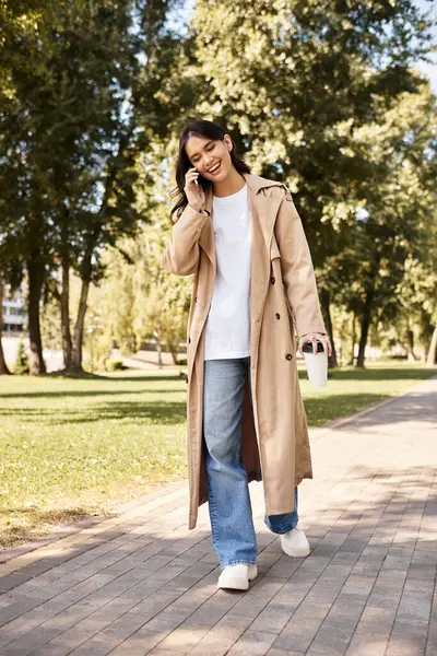 A young woman in warm clothes strolls through a scenic park, happily chatting on her phone. — Stock Photo