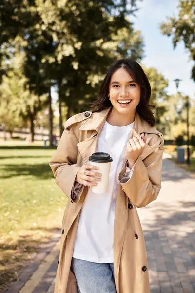 A young woman in cozy autumn attire radiates happiness while holding a warm drink in the park. — Stock Photo