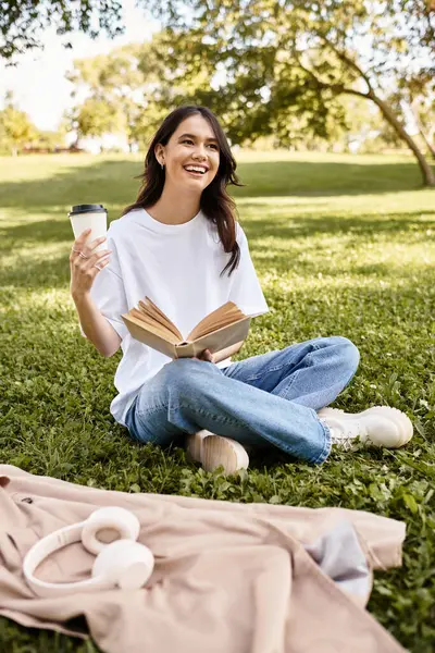 A smiling woman sits on the grass, sipping coffee while reading her book in a tranquil autumn park. — Stock Photo