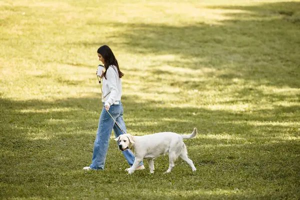 A young woman in warm autumn attire happily walks her dog through a serene park setting. — Stock Photo