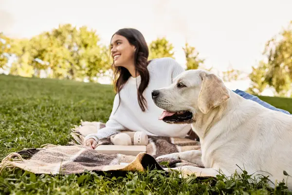 A young woman sits on a blanket in a sunlit park, smiling as she relaxes with her dog. — Stock Photo