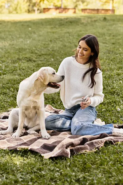 A young woman in cozy autumn attire smiles while playing with her dog on a picnic blanket. — Stock Photo