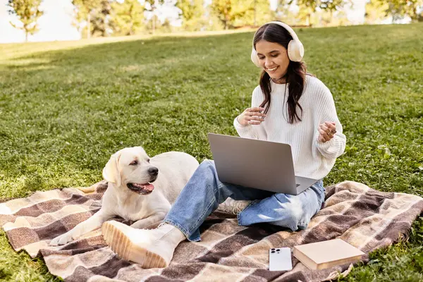 A young woman in warm attire sits on a blanket, joyfully using her laptop with a dog nearby. — Stock Photo