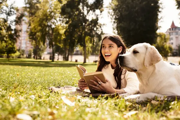 A young woman in cozy autumn attire sits on grass, happily reading with her dog beside her. — Stock Photo