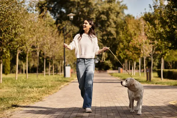 Wearing warm attire, a young woman joyfully strolls with her dog through a sunny park in autumn. — Stock Photo