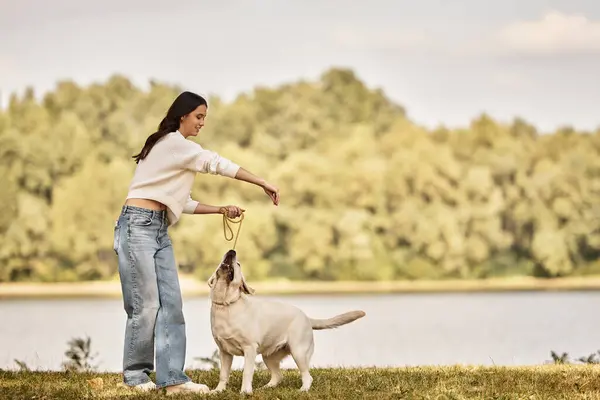 Eine junge Frau in kuscheliger Herbstkleidung spielt fröhlich mit ihrem Hund in der Nähe eines ruhigen Sees. — Stockfoto