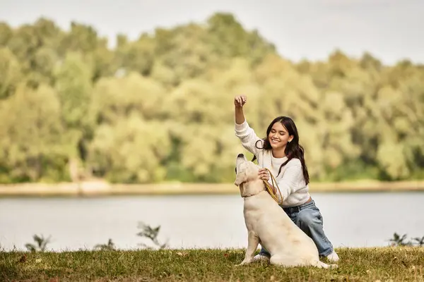 A young woman dressed in cozy autumn attire interacts joyfully with her dog near a serene lake. — Stock Photo