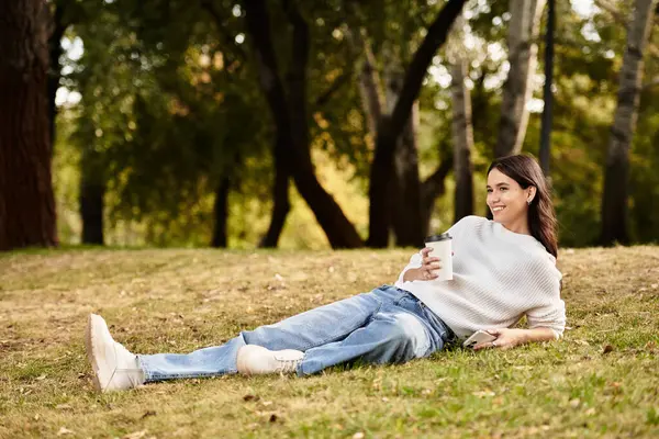 A young woman lounges in the park, sipping a warm drink while surrounded by autumn foliage. — Stock Photo