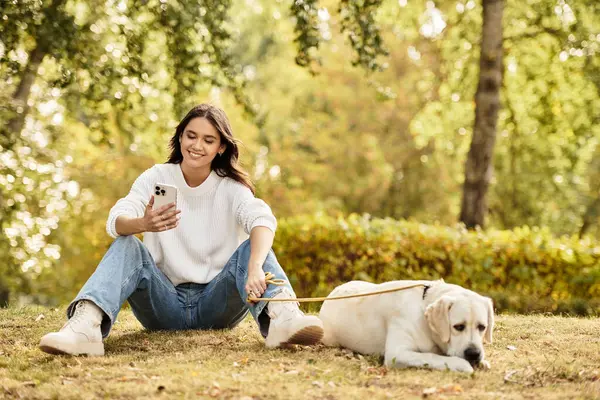 A beautiful young woman relaxes in the park, checking her phone beside her dog on a warm autumn day. — Stock Photo