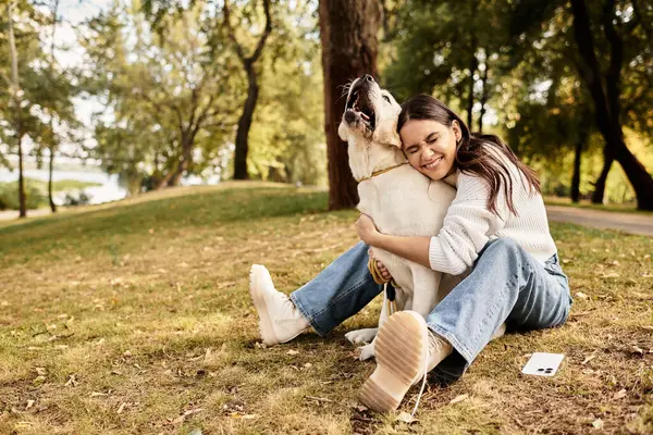 A delighted woman embraces her dog while sitting on the grass in a vibrant autumn park. — Stock Photo