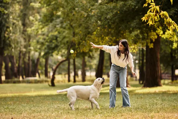 A young woman in cozy autumn clothes happily tosses a ball for her dog in a park. — Stock Photo