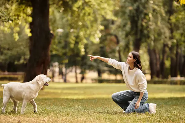 A beautiful woman in warm autumn attire kneels in a vibrant park, joyfully interacting with her dog. — Stock Photo