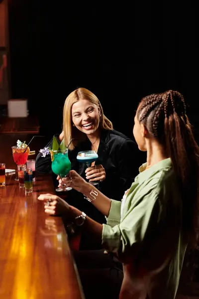 Two diverse colleagues share laughter while savoring colorful drinks at an energetic celebration. — Stock Photo