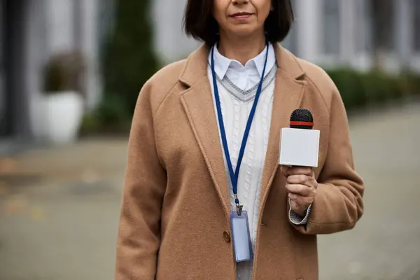 Un giornalista con un cappotto beige tiene un microfono, pronto a condividere le ultime notizie in un ambiente cittadino occupato. — Foto stock
