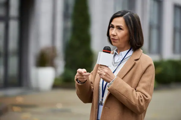 A multiracial female journalist in a beige coat speaks confidently into a microphone during a news report. — Stock Photo