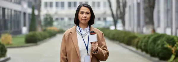Una donna multirazziale in un cappotto beige intervista all'aperto, tenendo un microfono in un cortile della città. — Foto stock
