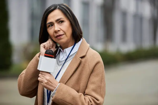 Una giornalista multirazziale femminile tiene un microfono e parla pensieroso mentre è all'aperto, vestita elegantemente. — Foto stock