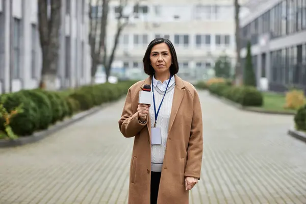 Un giornalista multirazziale con un cappotto beige tiene un microfono mentre riferisce in diretta in un ambiente urbano. — Foto stock
