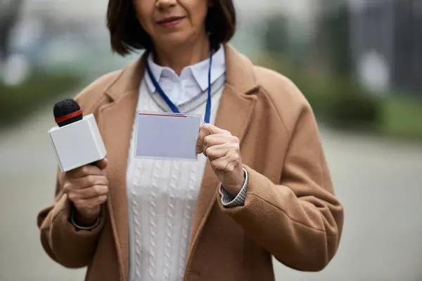 Una periodista multirracial con un abrigo beige sostiene un micrófono y muestra su insignia de prensa - foto de stock
