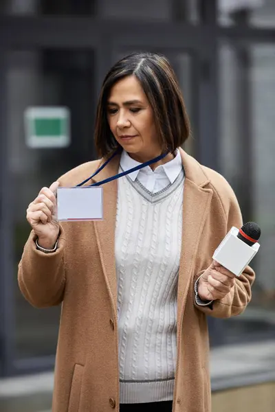 Eine multiethnische Journalistin steht draußen, hält ein Mikrofon in der Hand und zeigt ihr Presseabzeichen, bereit zu berichten. — Stockfoto