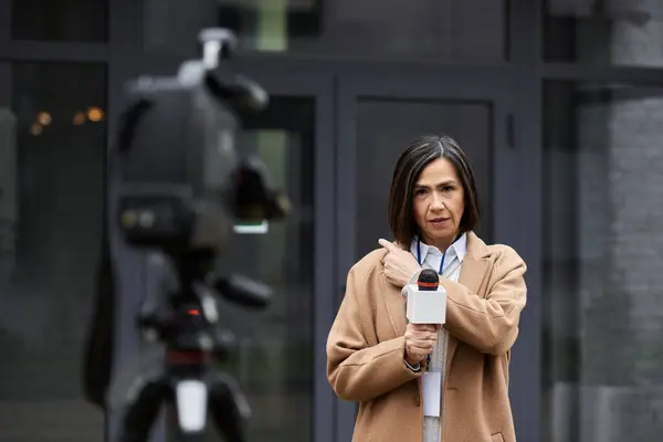 A journalist wearing a beige coat is reporting live, holding a microphone and engaging with cameras outside. — Stock Photo
