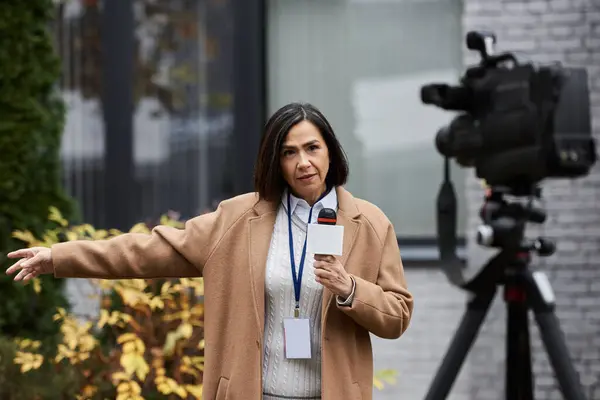 Una giornalista multirazziale con un cappotto beige sta in piedi con fiducia, tenendo un microfono e trasmettendo in diretta. — Foto stock