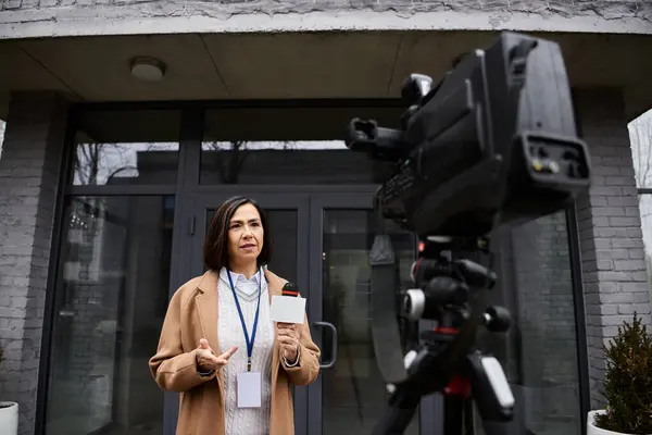 Una donna multirazziale in un cappotto beige si erge con fiducia con un microfono, pronto a riferire le notizie. — Foto stock