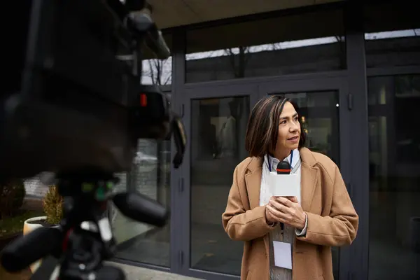 Un giornalista con un cappotto beige tiene un microfono, preparandosi a riferire notizie fuori da un edificio elegante. — Foto stock