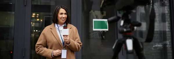 Una giornalista multirazziale femminile tiene un microfono mentre consegna aggiornamenti all'aperto in un cappotto beige. — Foto stock