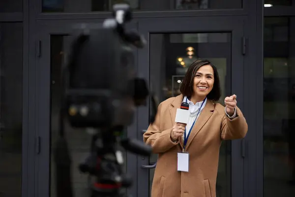 Una mujer multirracial con un abrigo beige entrevista con entusiasmo fuera, capturando el momento con su micrófono. - foto de stock