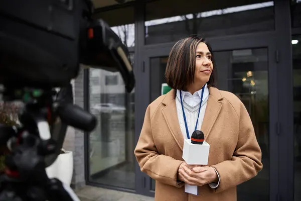 A multiracial journalist in a beige coat prepares to deliver news while holding a microphone outside a building. — Stock Photo