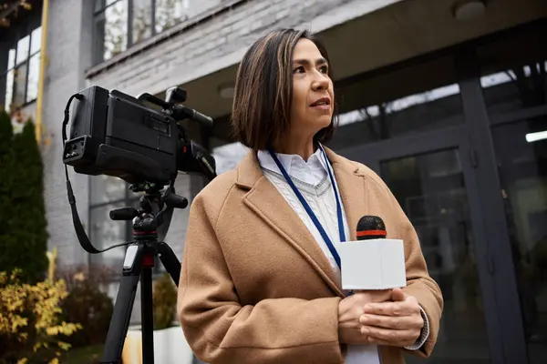 A multiracial female journalist in a beige coat stands confidently outside, preparing to report news. — Stock Photo