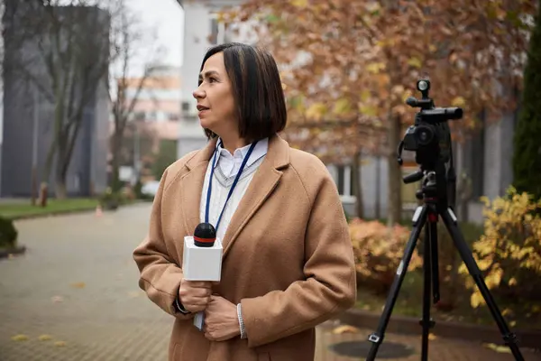 Un giornalista multirazziale che indossa un cappotto beige si distingue all'aperto con un microfono, preparandosi a condividere aggiornamenti sulle notizie. — Foto stock