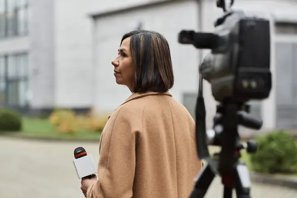 Un giornalista multirazziale con un cappotto beige tiene un microfono, che si prepara a riferire sugli eventi in corso all'aperto. — Foto stock