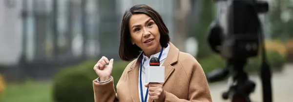 Una giornalista multirazziale con un cappotto beige riporta appassionatamente le notizie all'aperto mentre tiene in mano un microfono. — Foto stock