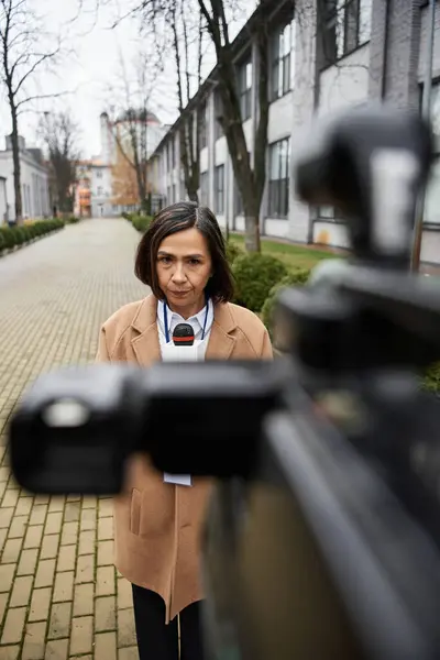 Una giornalista multirazziale con un cappotto beige si alza con fiducia, microfono in mano, pronta a condividere le notizie. — Foto stock