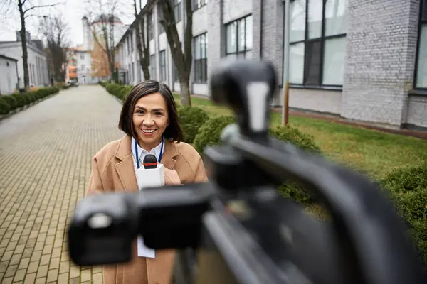 Uma jornalista multirracial se destaca ao ar livre, sorrindo e relatando com um microfone em um ambiente urbano. — Fotografia de Stock