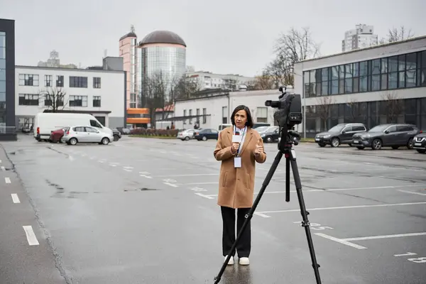 Una giornalista multirazziale in cappotto beige è pronta con un microfono in un ambiente urbano. — Foto stock