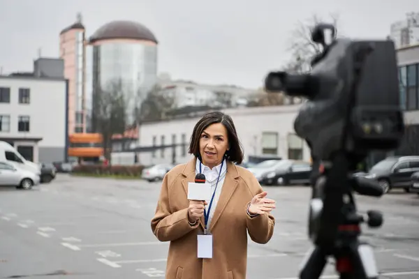 Um jornalista multirracial com um casaco bege está em um estacionamento, entregando com confiança uma reportagem. — Fotografia de Stock