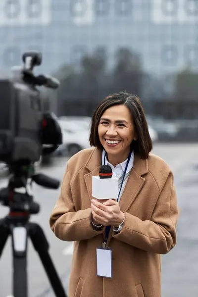 A multiracial journalist in a beige coat eagerly conducts an outdoor news report, microphone in hand. — Stock Photo