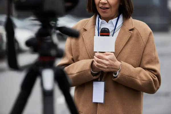 A multiracial female journalist in a beige coat holds a microphone while preparing to report from a busy street. — Stock Photo