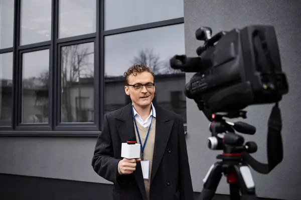 Un giornalista professionista in occhiali parla in un microfono mentre si trova vicino a una telecamera all'aperto. — Foto stock