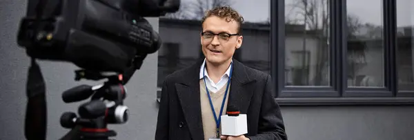 Un giornalista con gli occhiali e il cappotto parla in un microfono, condividendo importanti aggiornamenti all'esterno. — Foto stock